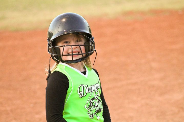 Little girl in baseball helmet and green jersey smiling on the field.